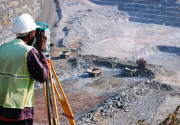 An African man, locally employed as a surveyor at an open-pit copper mine in Zambia, peers through his survey instrument.  This work records the daily changes in the open-pit, and help guide mining activities to the engineer's plans.